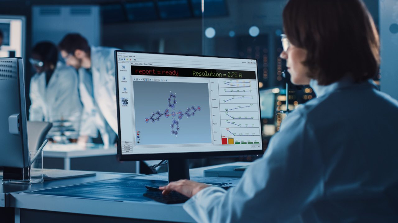 Over the Shoulder Shot: Female IT Scientist Uses Computer Showing System Monitoring and Controlling Program. In the Background Technology Development Laboratory with Scientists, Engineers Working