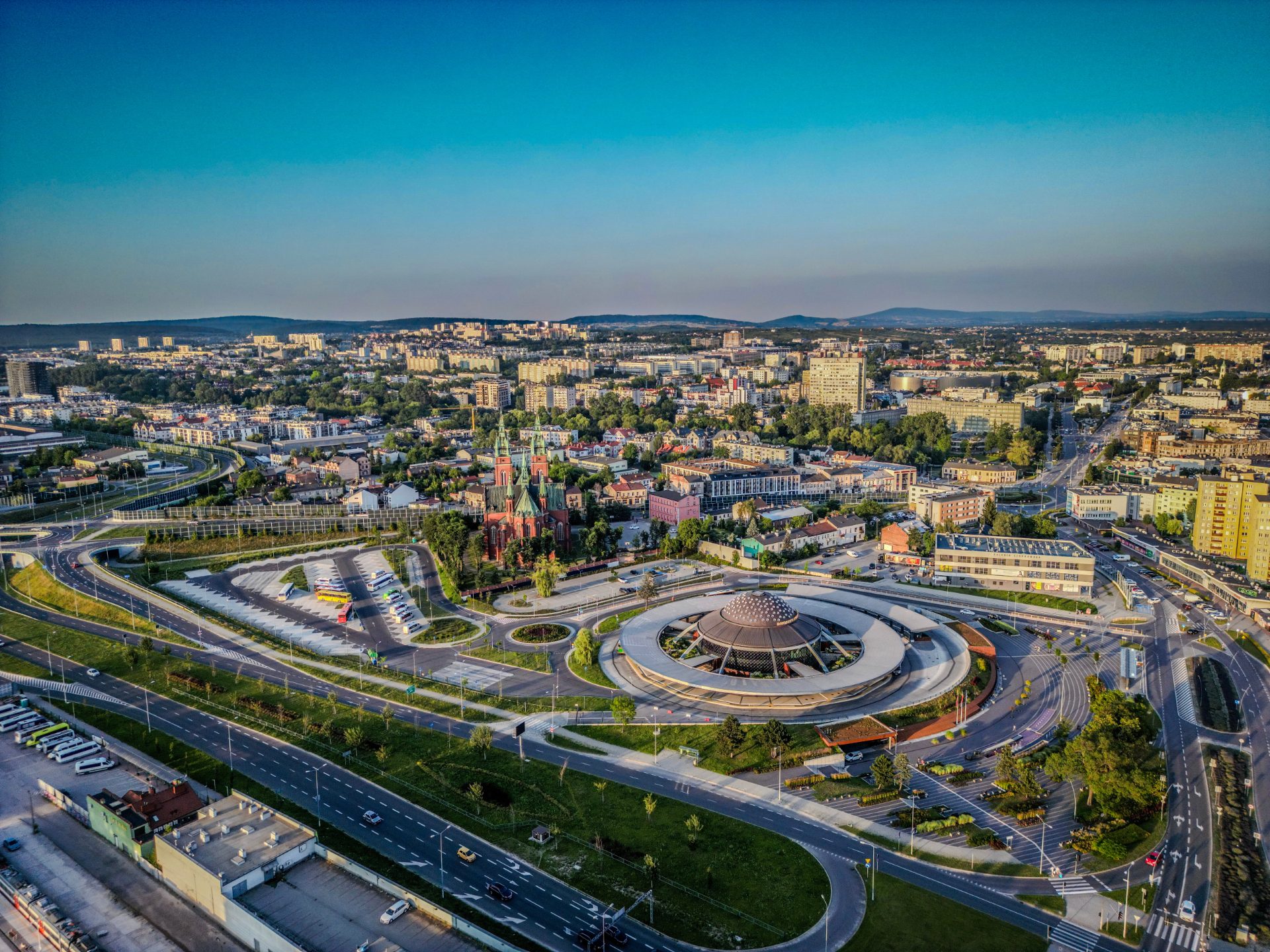 Ariel view of kielce poland, lots of buildings and roads can be seen with green fields and small hills in the distance under a bright blue sky.