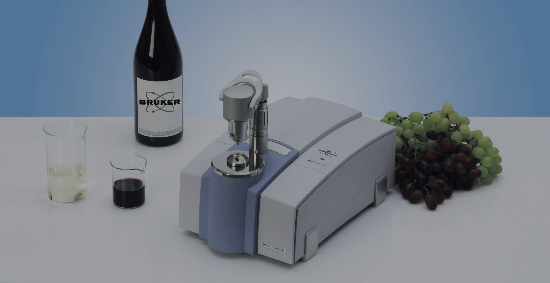  The image shows a Bruker ALPHA II FTIR spectrometer (wine analyzer) on a table alongside various items related to wine analysis. There are two beakers containing white and red wine samples, a bottle of wine labeled "Bruker," and a bunch of green and red grapes. The spectrometer has a platinum ATR accessory attached, suggesting its use for analyzing wine samples. The scene highlights the use of scientific instruments in food and beverage quality control, particularly in wine testing. The background is a light blue gradient, giving the image a clean, professional appearance.