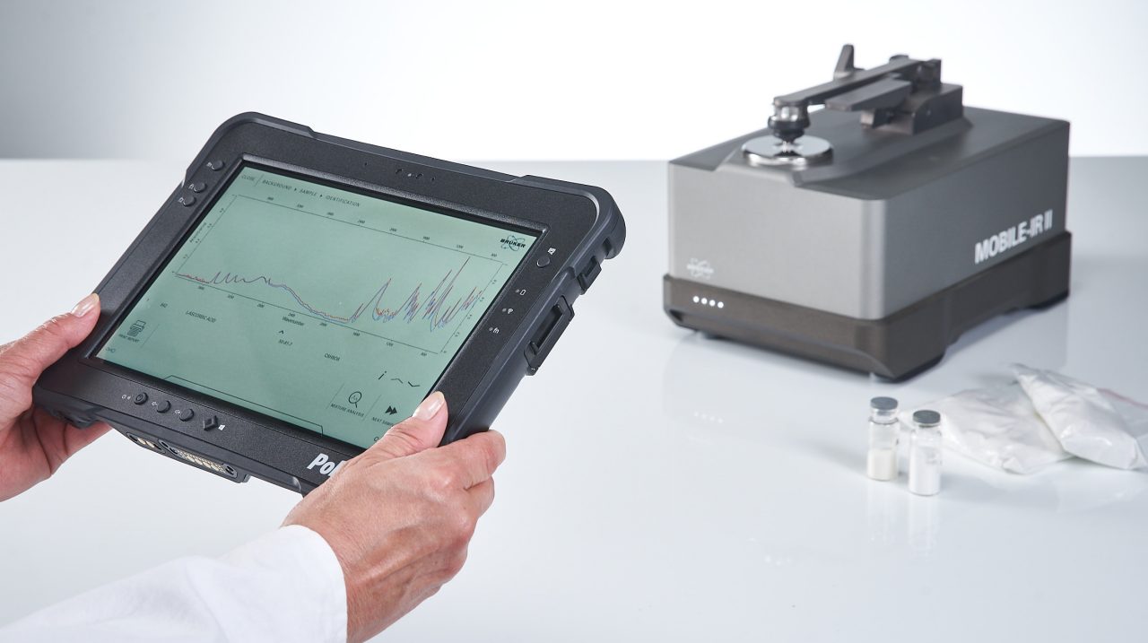 La personne tient la tablette dans les mains. La tablette affiche une mesure IRTF réussie. Le spectromètre IRTF mobile est montré flou.