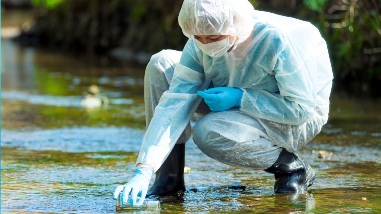 A scientist wearing a protective suit is taking a water sample from a river.