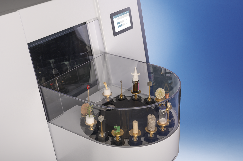 Rendering showing the X4 POSEIDON with the optional 15-position sample changer attached to its front. The sample holder contains a range of different samples, some stacked on one another. The sample holder sits on the front of the machine.