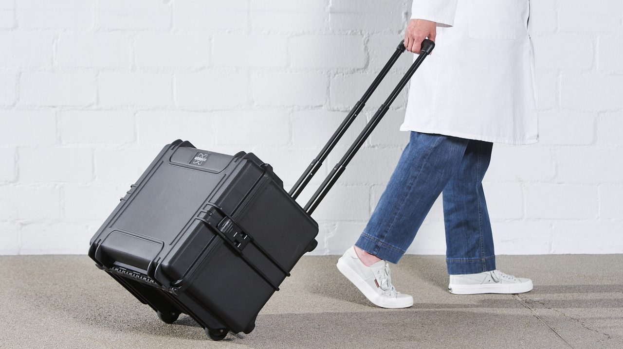 A carry case with extended handle is pulled by a person in a lab coat.